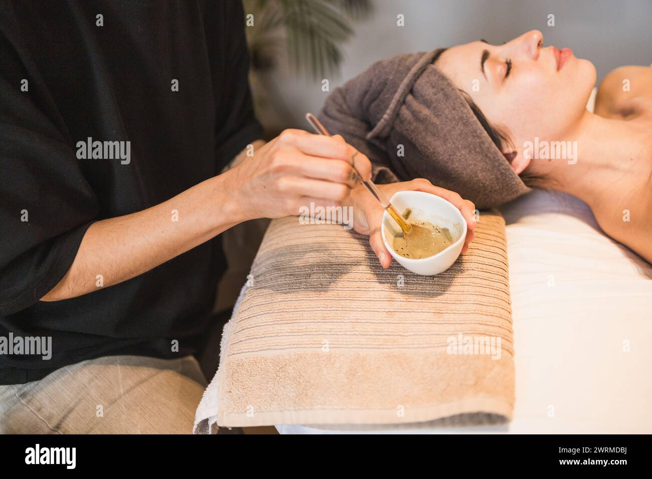 Eine erfahrene Kosmetikerin bereitet einer entspannten Kundin in einer ruhigen Wellness-Spa-Umgebung sanft eine grüne Gesichtsmaske vor Stockfoto