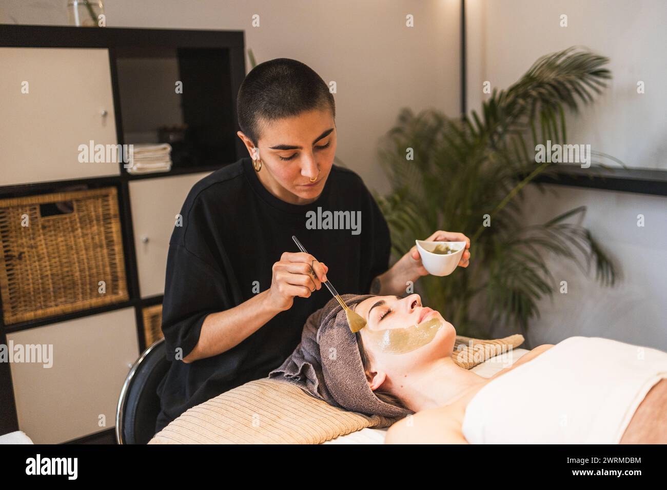 Eine gelernte Kosmetikerin wendet eine grüne Gesichtsmaske sanft auf eine entspannte weibliche Kundin in einer ruhigen Wellness-Spa-Umgebung an Stockfoto