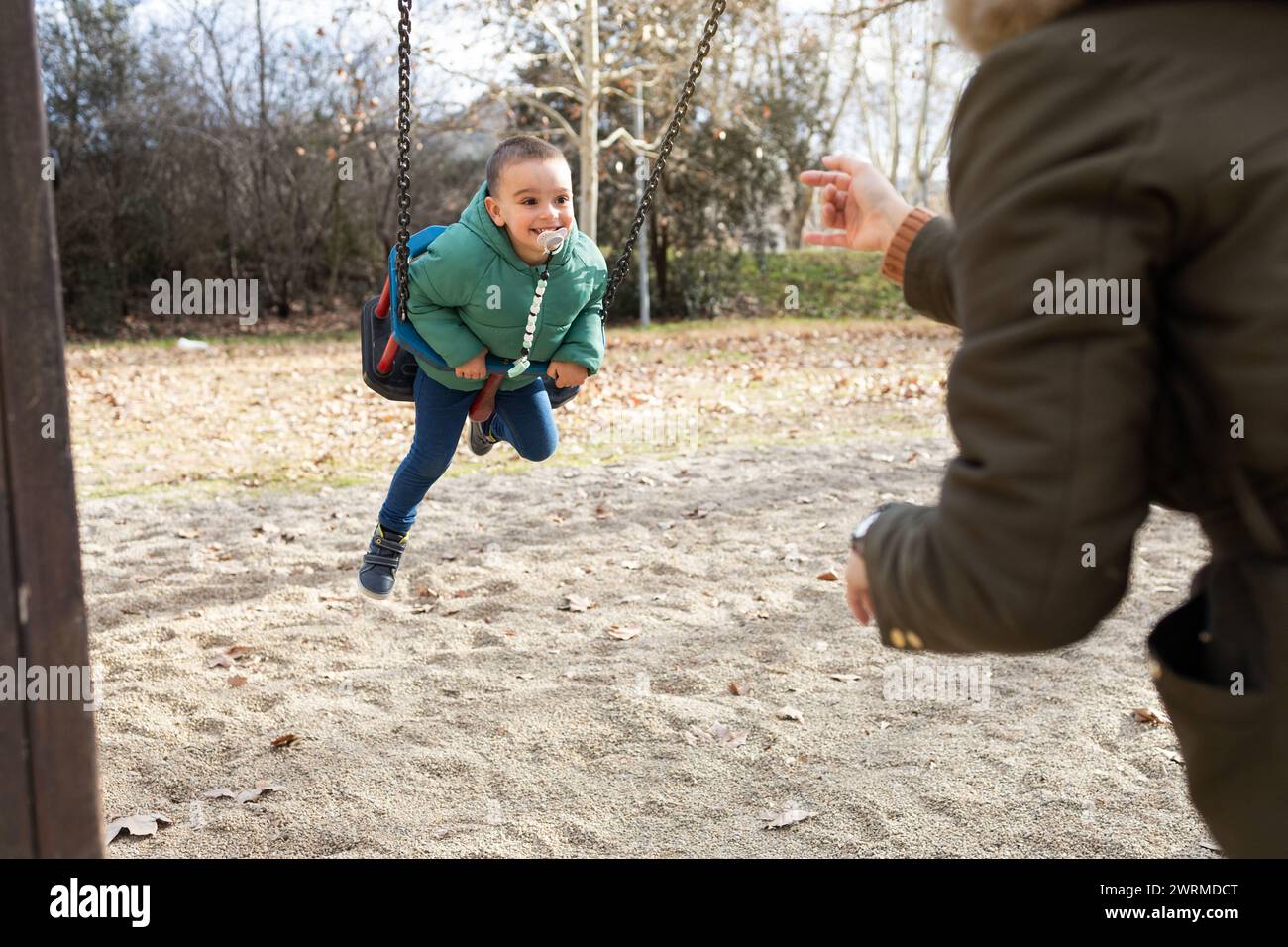 Ein verspielter Moment, in dem ein kleines Kind kichert, während es sich in einem sonnigen Park auf eine ausgebreitete anonyme Hand schwingt Stockfoto