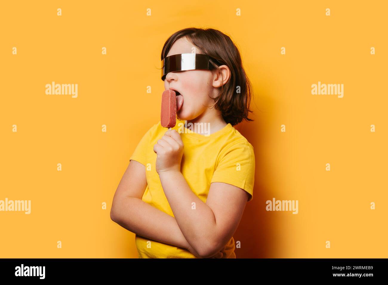 Ein Kind in Sonnenbrille und gelbem Hemd leckt ein Eis vor orangefarbenem Hintergrund und weckt Sommerfreude Stockfoto