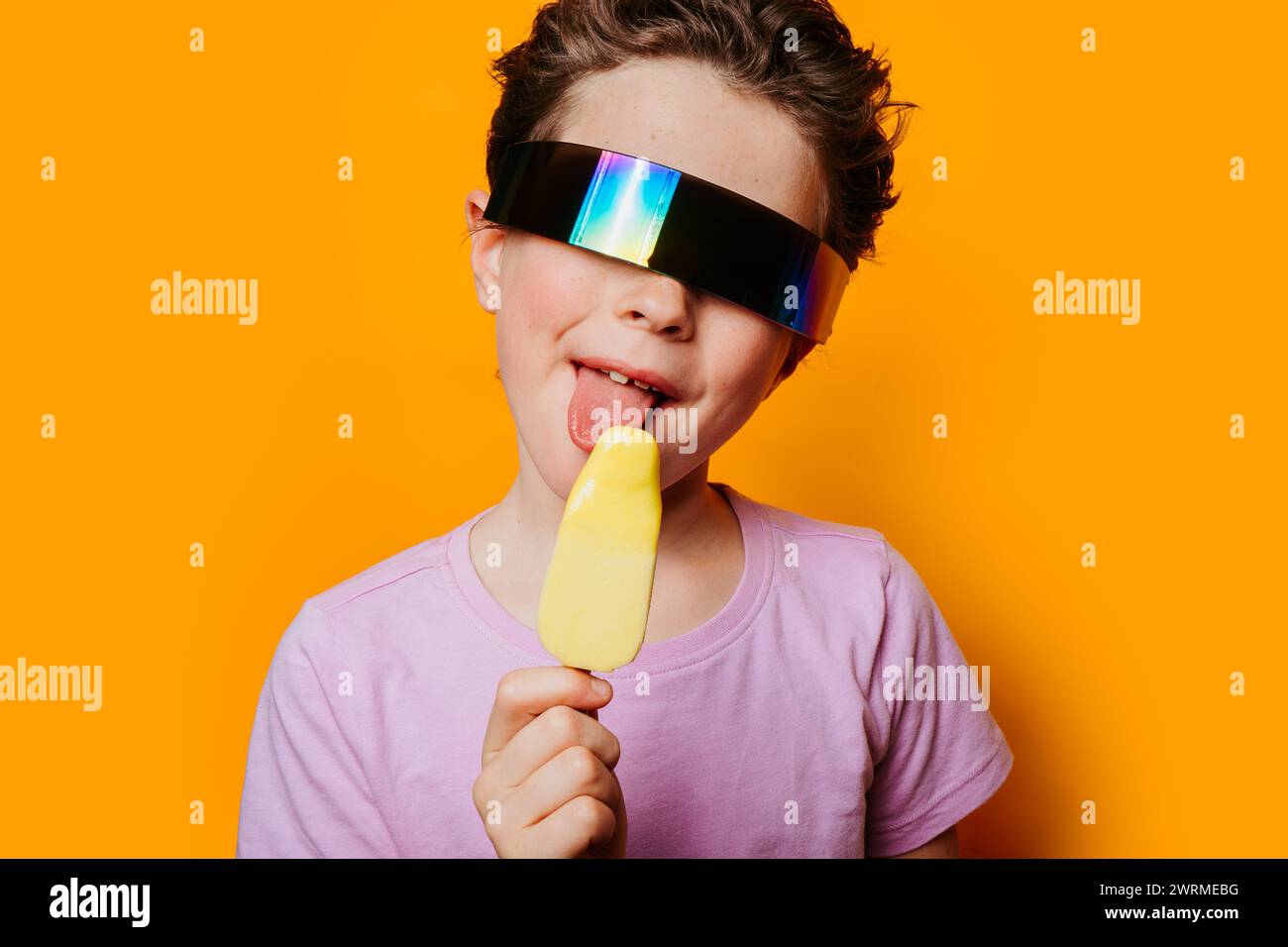 Ein kleines Kind mit reflektierender, futuristischer Sonnenbrille genießt ein gelbes Eis vor einem leuchtend orangefarbenen Hintergrund, das ein Gefühl von Sommerspaß vermittelt Stockfoto