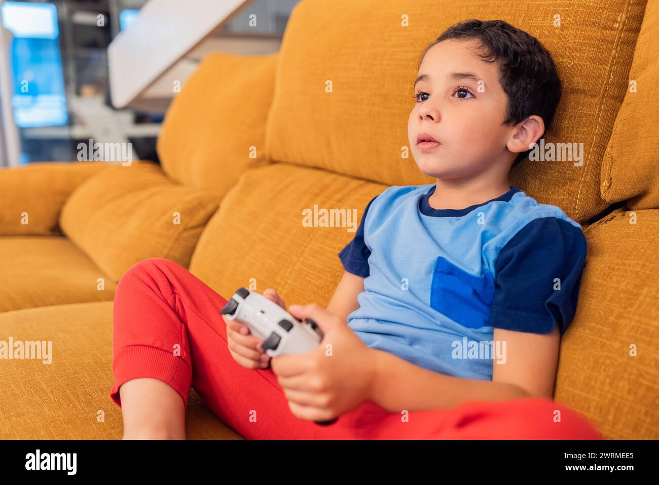 Junge, der mit Videospielen auf einer senffarbenen Couch sitzt, mit einem Controller in den Händen Stockfoto