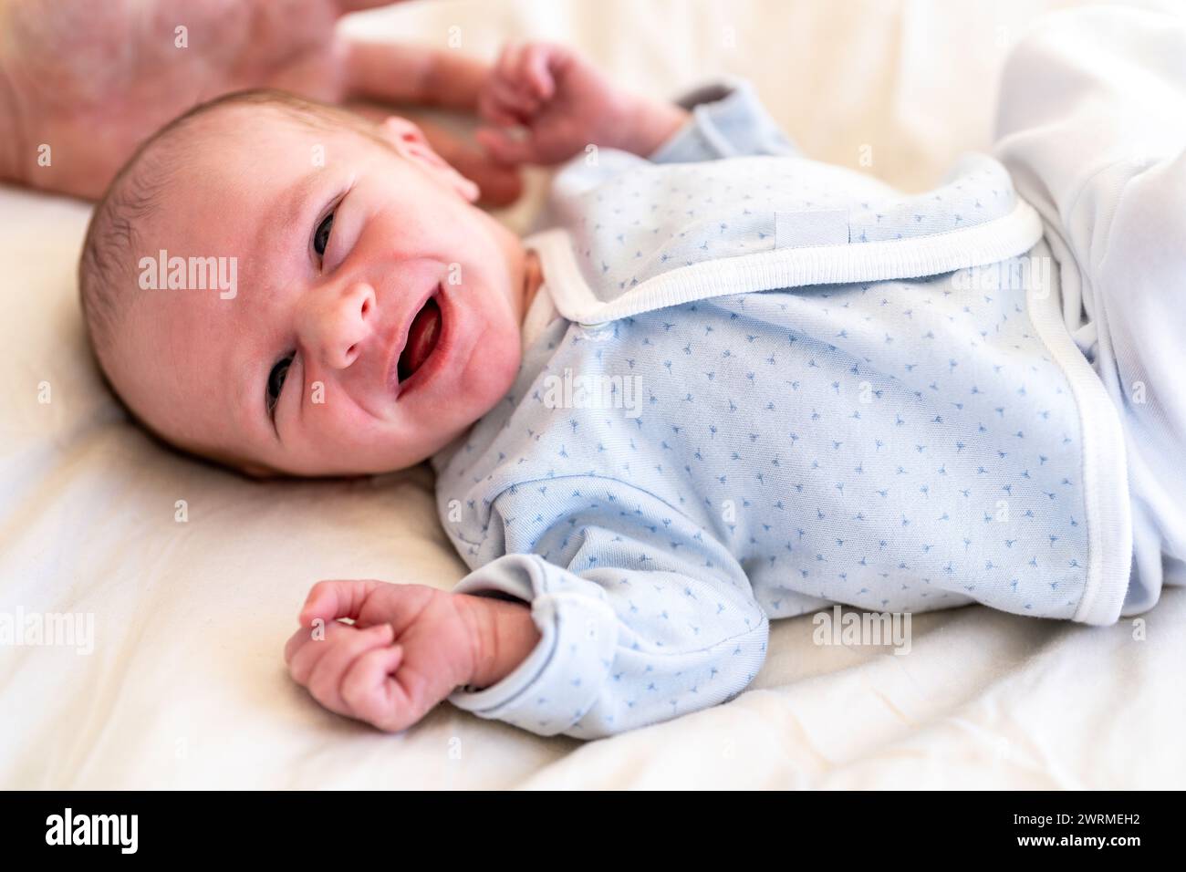 Ein waches Neugeborenes in einem gemusterten Einteiler liegt auf einer weichen Oberfläche und blickt mit neugierigen Augen und einem teilweise offenen Mund Stockfoto
