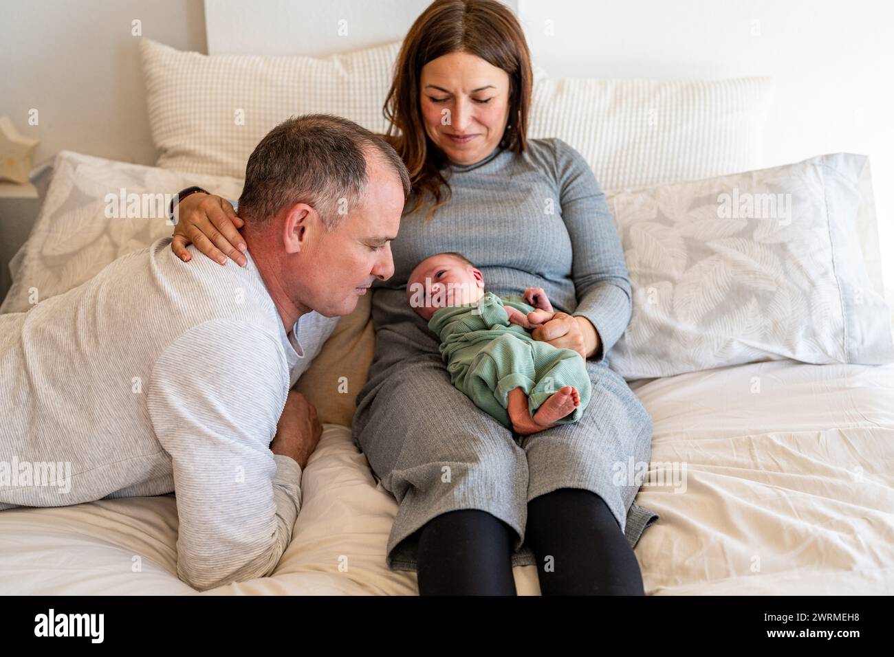 Ein Paar sitzt auf dem Bett und liebt ihr friedliches, schlafendes Neugeborenes, das im Schoß der Mutter liegt, und teilt einen ruhigen Familienmoment Stockfoto