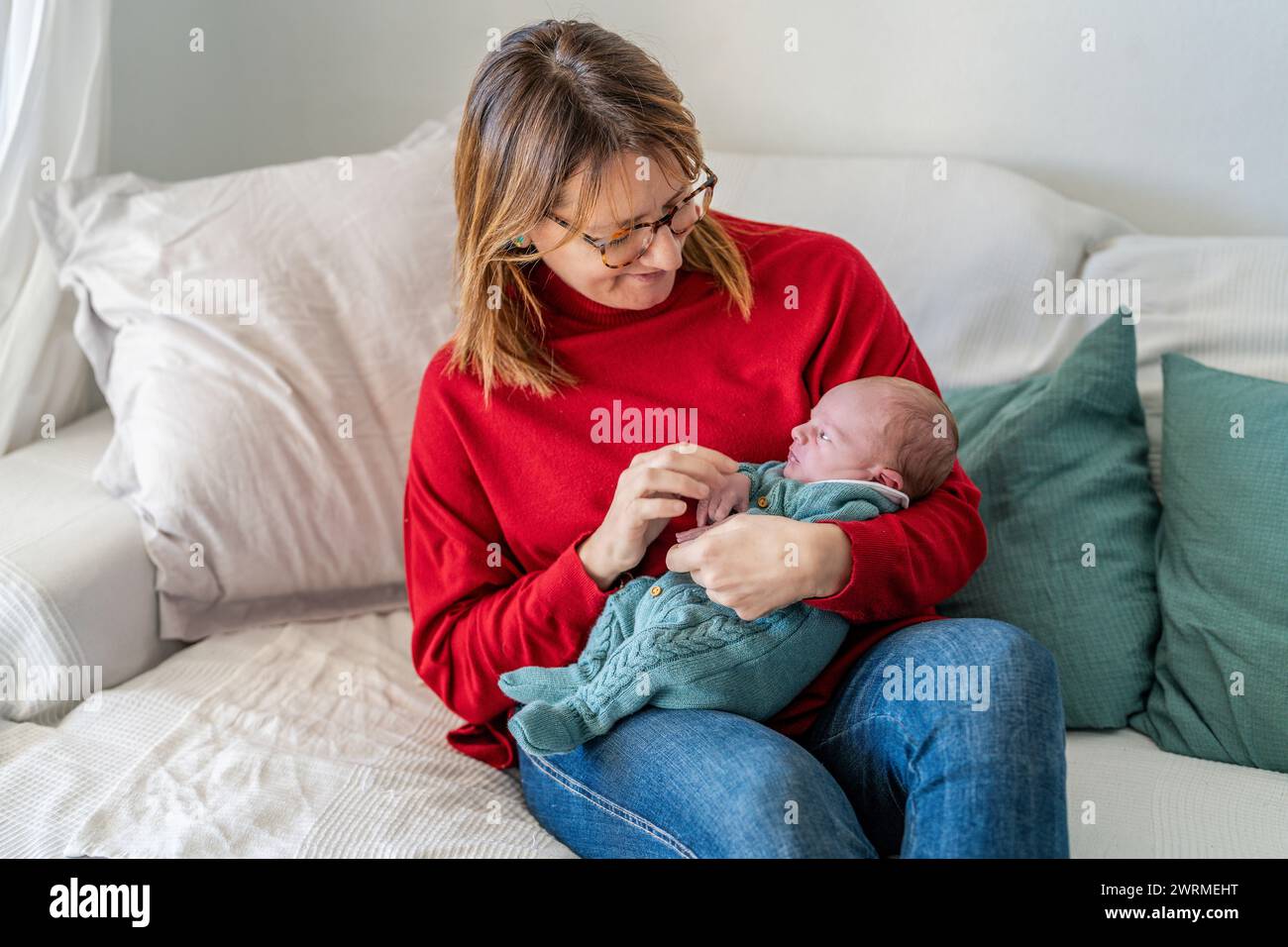 Eine Mutter in einem leuchtend roten Pullover kuschelt ihr Neugeborenes liebevoll und genießt die kostbaren Momente der frühen Mutterschaft zu Hause Stockfoto