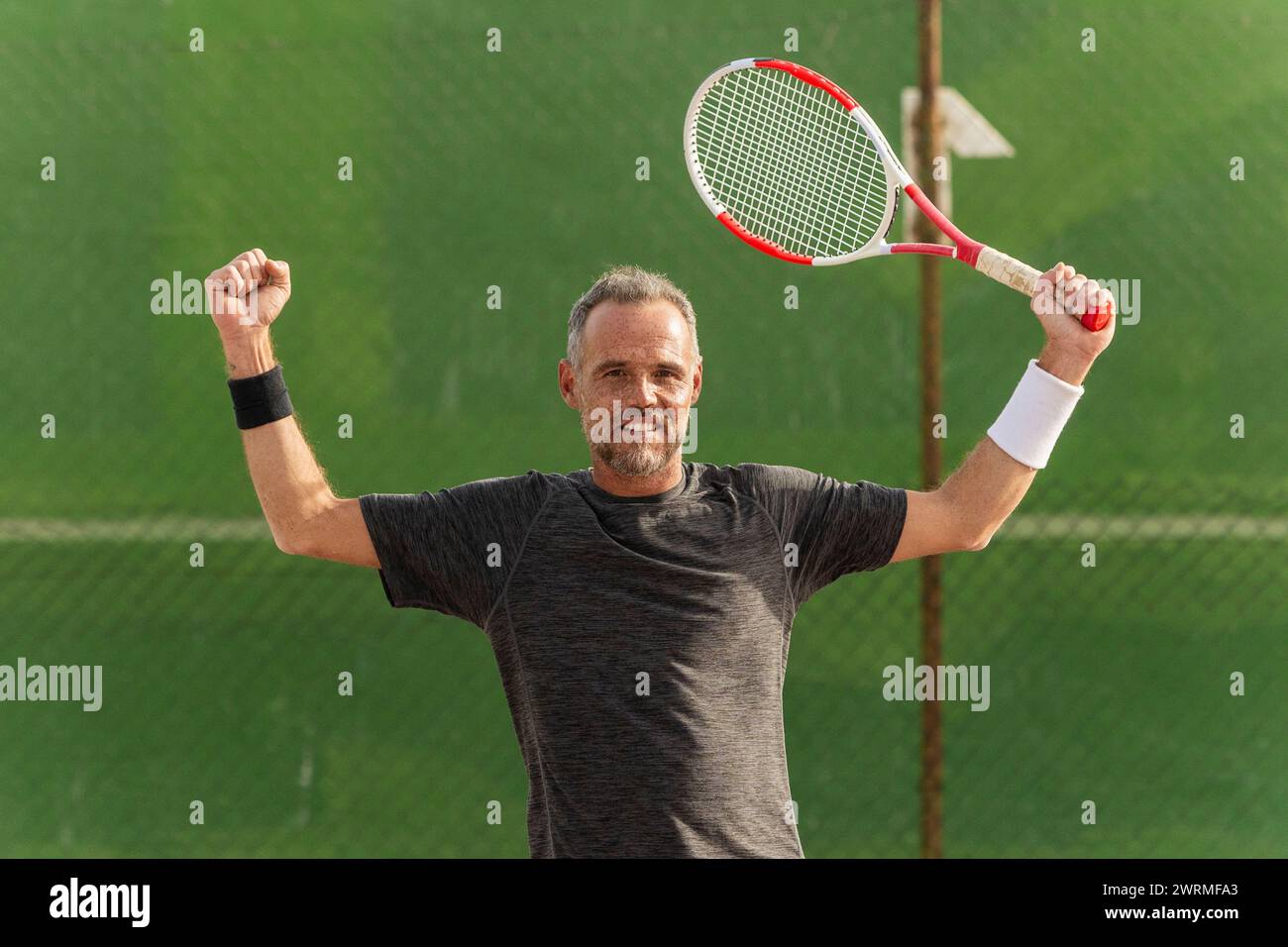 Ein fröhlicher Tennisspieler feiert einen Sieg auf dem Tennisplatz, hält seinen Schläger triumphierend und blickt mit einem stolzen Lächeln in die Kamera. Stockfoto