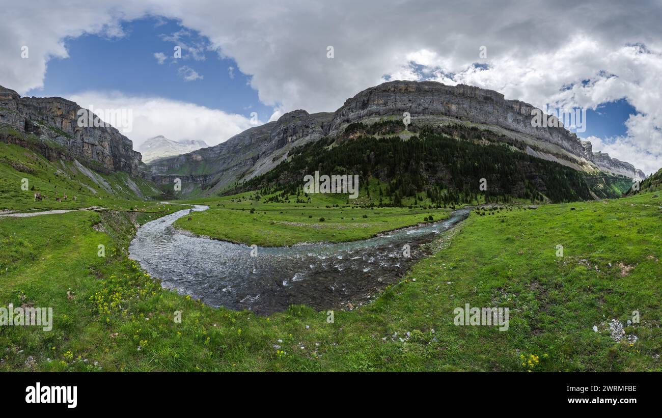 Ein Panoramablick auf das ruhige Valle de Ordesa, mit einem sich windenden Fluss, der durch üppige Wiesen und hoch aufragende Klippen unter einem dynamischen Himmel führt Stockfoto