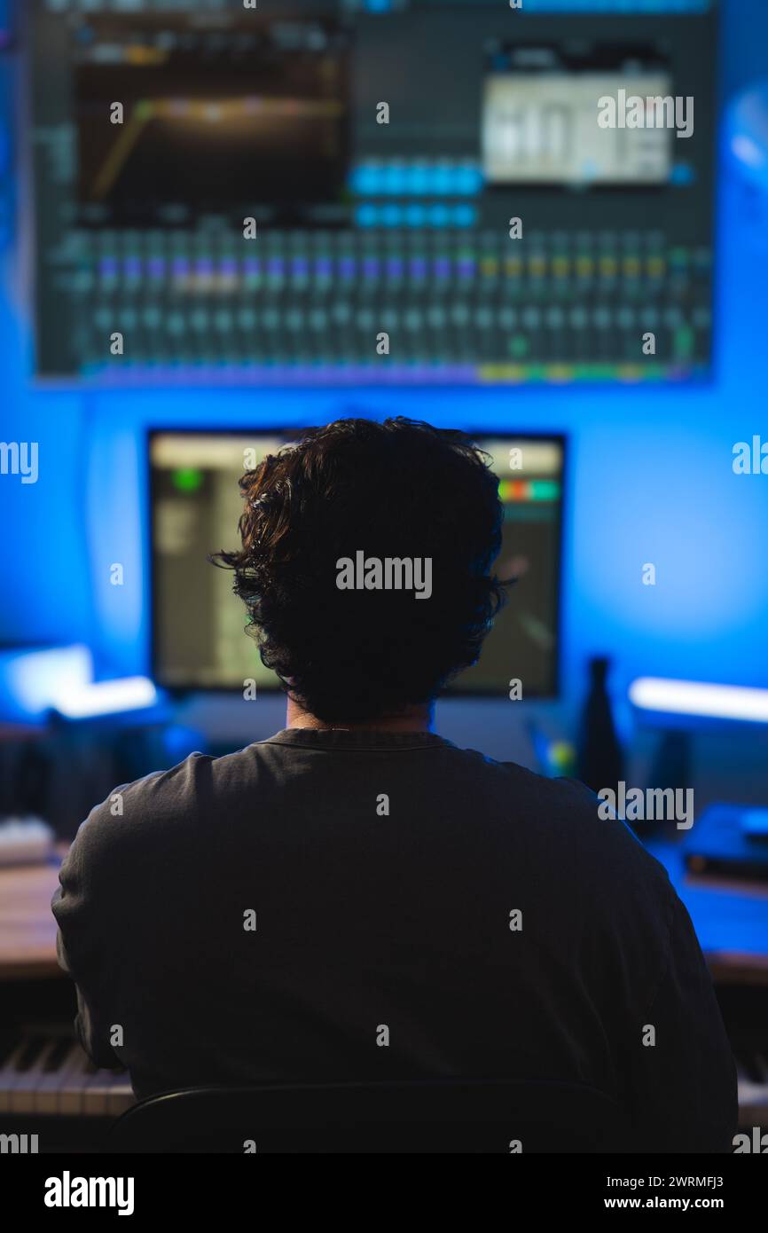 Ein Musikproduzent konzentriert sich auf das Mischen von Titeln in einem schwach beleuchteten Tonstudio, umgeben von professioneller Audioausrüstung und blauer Umgebungsbeleuchtung. Stockfoto