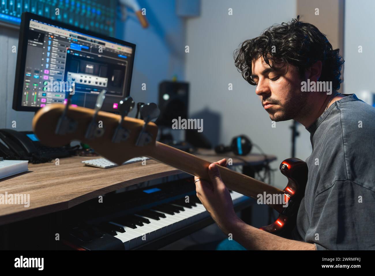 Ein fokussierter männlicher Musiker stimmt eine Bassgitarre vor dem Hintergrund einer digitalen Klangbearbeitung in einer modernen Studio-Umgebung ab. Stockfoto