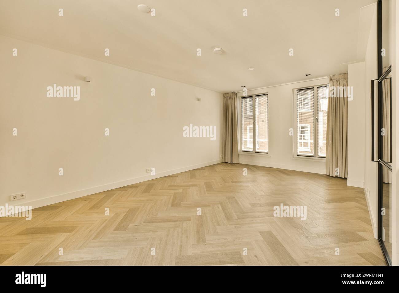 Ein leeres Wohnzimmer mit Sonnenlicht, das durch drei große Fenster strömt und sich von Holzböden mit Fischgrätenmuster und weißen Wänden reflektiert. Stockfoto