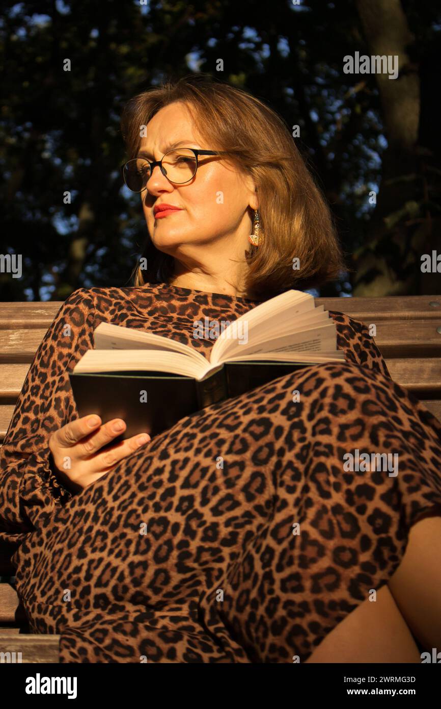 Frau im Leopardenkleid mit offenem Buch im Park. Frau in Brille liest Buch auf der Bank. Lifestyle in der Großstadt. Literaturkonzept. Wunderschöner Lehrer. Stockfoto