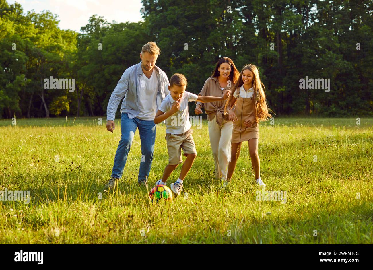 Fröhlich lächelnde vierköpfige Familie, die gemeinsam im Sommerpark auf den Ball läuft. Stockfoto