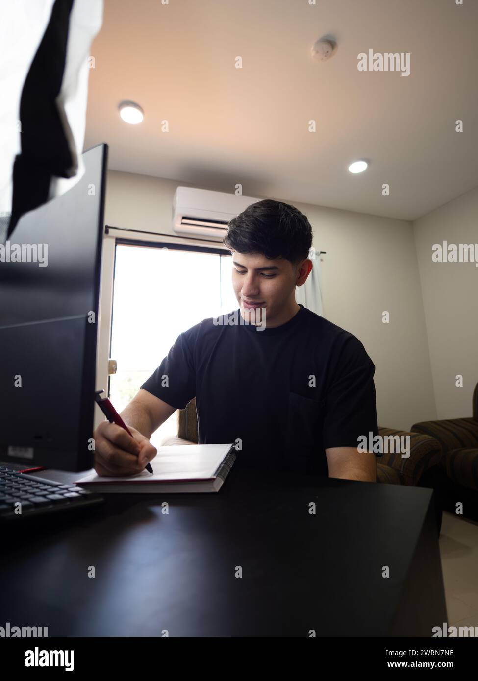 Bild eines lächelnden, glücklichen jungen Mannes, der zu Hause an seinem Schreibtisch sitzt, mit einem Computer vor ihm Stockfoto
