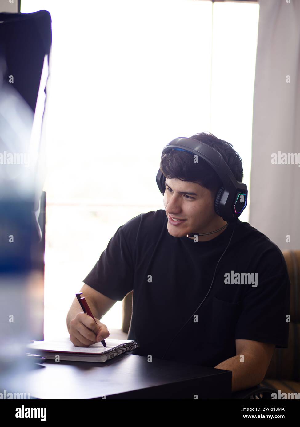 Ein lächelnder junger Mann schreibt in einem Notebook mit einem Stift, mit Kopfhörern vor seinem Computer, taucht in Arbeit oder Studium ein Stockfoto