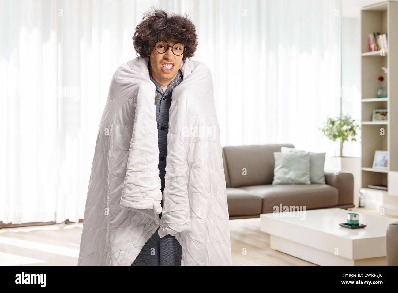 Ein junger Mann, in eine Decke gewickelt, steht in einem Wohnzimmer und fühlt sich krank Stockfoto