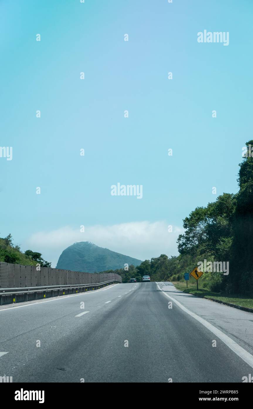 Blick auf KM 5 der Via Lagos Highway mit einem großen Berg, der von dichter Atlantikwaldvegetation und Wolken im Hintergrund unter dem klaren blauen Sommerhimmel bedeckt ist. Stockfoto