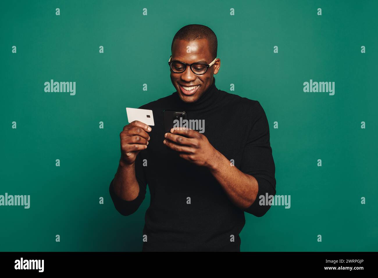 Der modische junge Mann, lässig gekleidet und mit Brille, steht vor einem grünen Hintergrund und lächelt, während er sein Smartphone zum Online-Shopping benutzt Stockfoto