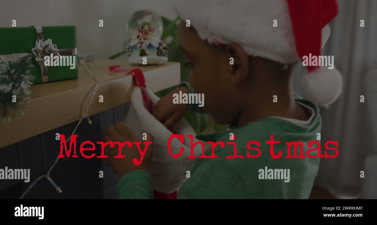 Bild von Merry christmas Text über dem afroamerikanischen Jungen, der weihnachtsmütze trägt Stockfoto