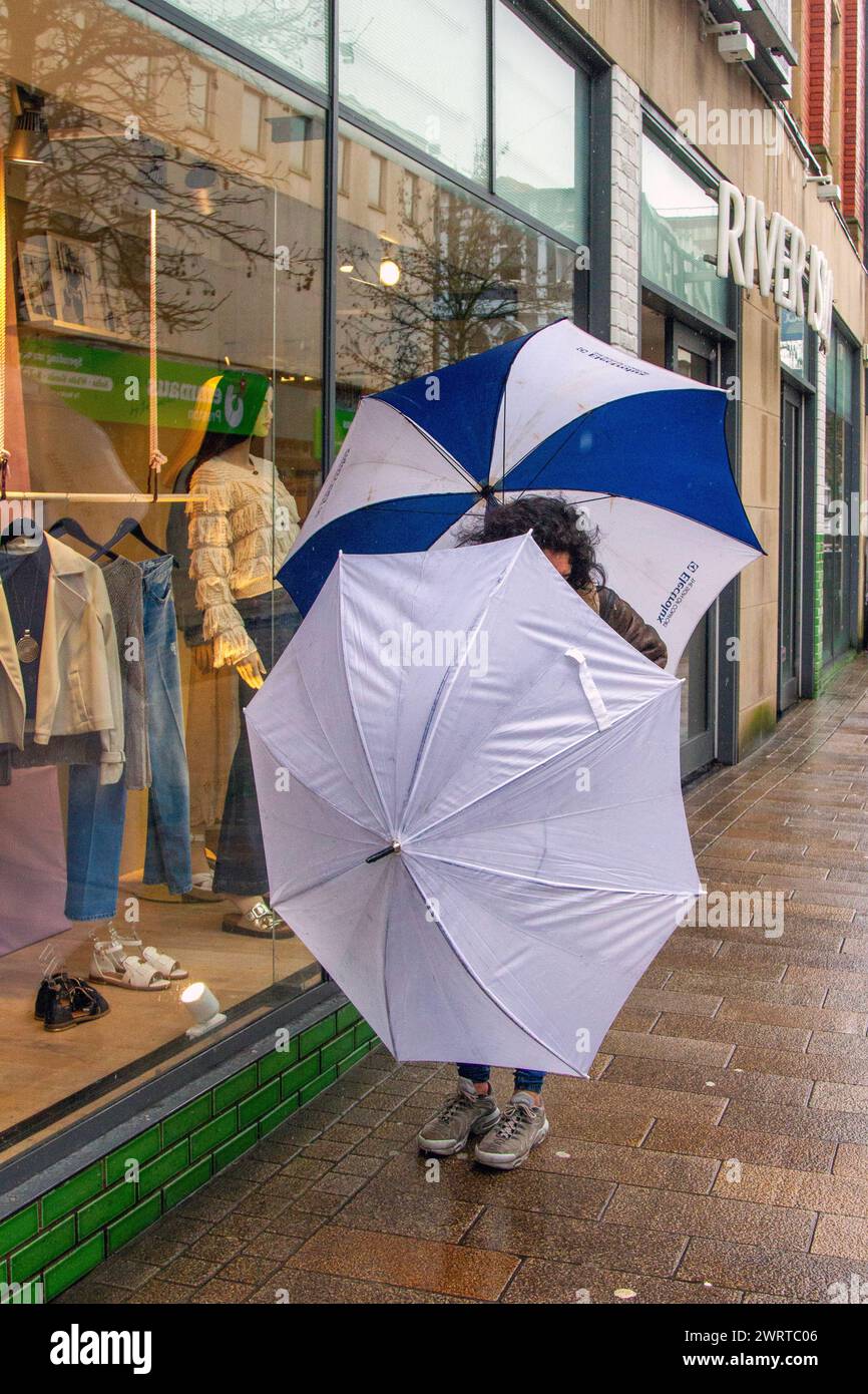 Frau, die zwei Regenschirme an einem böigen Tag in Preston, Lancashire, trägt. Wetter in Großbritannien. März 2024. Regnerischer Start in den Tag für Shopper im Stadtzentrum. Wind mit heftigeren Regenfällen. Credit; MediaWorldImages/AlamyLiveNews Stockfoto