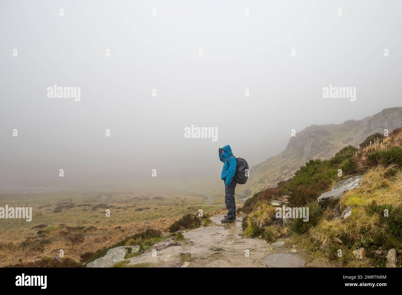 Seitenansicht eines Mannes, der im Regen auf einem Berg in Nebel schaut, ist wasserdicht gekleidet und trägt einen großen Rucksack auf dem Rücken. Stockfoto