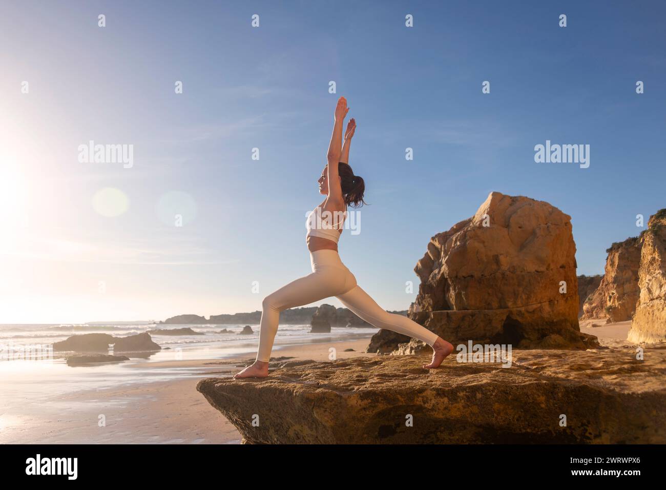 Frau, die auf Felsen am Meer steht und Yoga bei Sonnenaufgang praktiziert, Kriegerpositionen Stockfoto