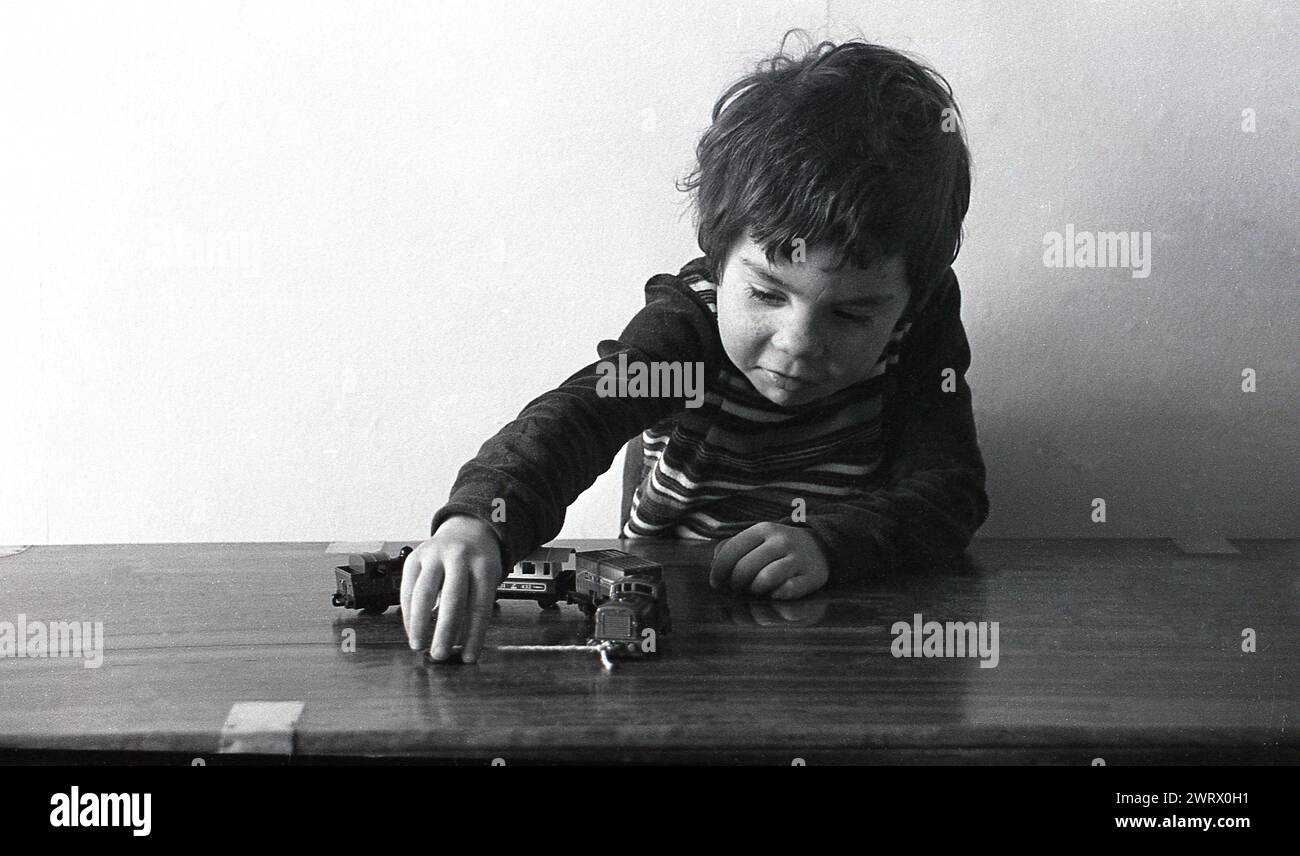 1970er Jahre, historisch, ein kleiner Junge, der mit Spielzeugzügen und Wagen auf einem Tisch spielt und die Schnur an der Vorderseite der Spielzeuglokomotive zieht, England, Großbritannien. Spielen ist ein wichtiger Teil des Lernens und der Entwicklung eines Kindes, bei dem es seine Fantasie erforscht und nutzt. Stockfoto
