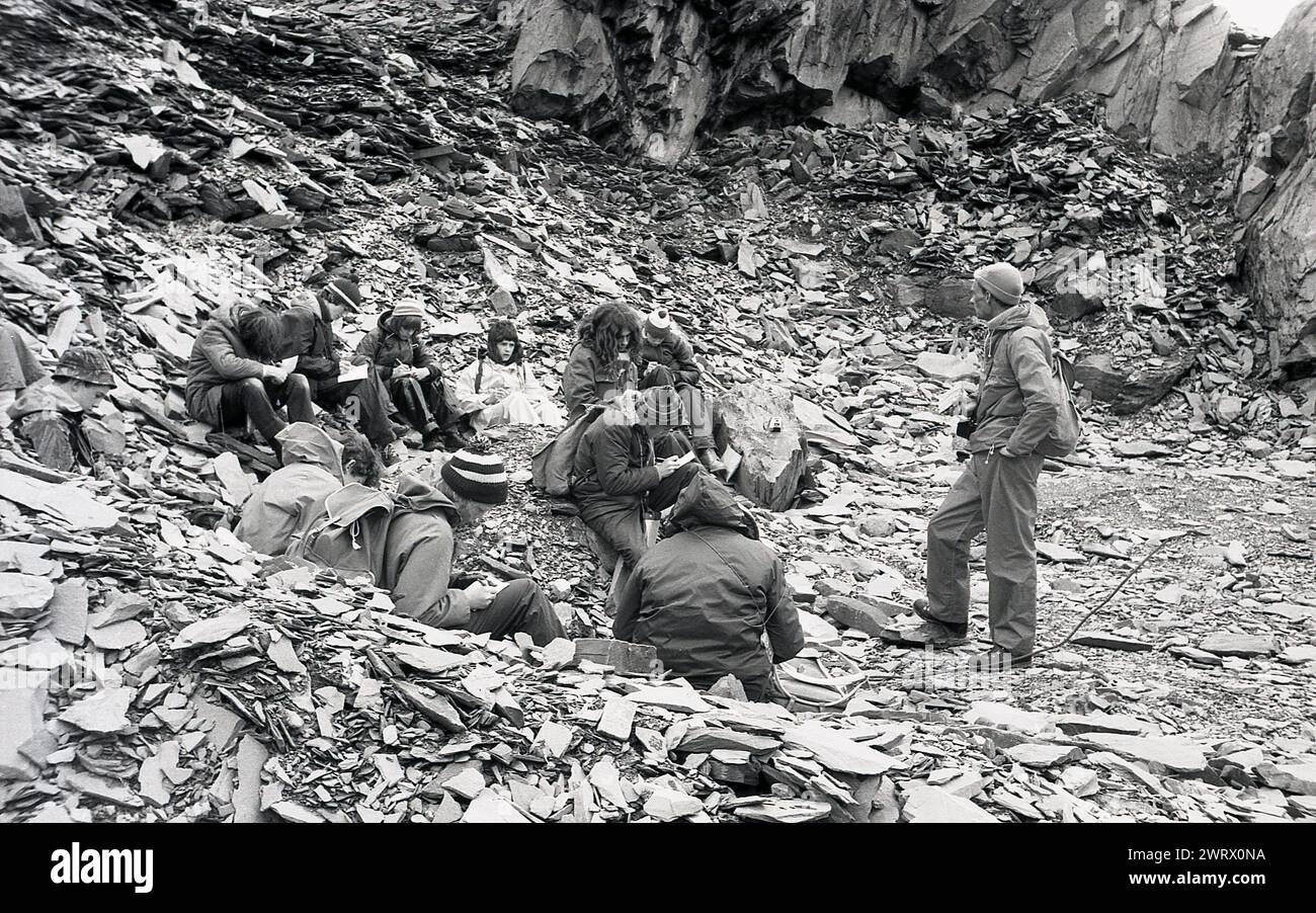 1970er Jahre, historisch, eine Gruppe junger Menschen mit begleitendem Erwachsenen, alle in Outdoor-Kleidung, auf dem Boden in einer Schiefergrube auf einem Hügel in der Nähe von Ambleside im Lake District, Cumbria, England, Großbritannien, während sie an einem Kurs nach außen teilnehmen. Die Felsen im Lake District umfassen eine Vielzahl verschiedener Arten, einschließlich des Schiefers, der hier zu sehen ist, sowie Sandstein, Kalkstein und vulkanisches Gestein. Stockfoto