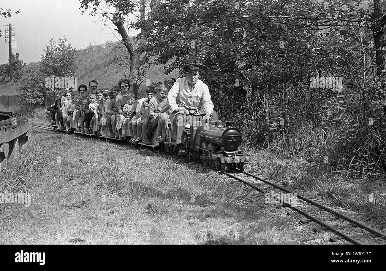 1970er Jahre, historisch, Erwachsene und Kinder fahren auf einer Miniatureisenbahn, England, Großbritannien, Zugführer im Overall und Hut, der die kleine Dampfeisenbahn auf der Außenbahn fährt. Stockfoto