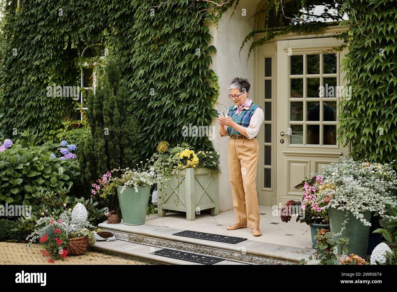 Raffinierte, fröhliche, reife Frau mit Brille, die ihr Handy in der Nähe ihres Hauses in England anschaut Stockfoto