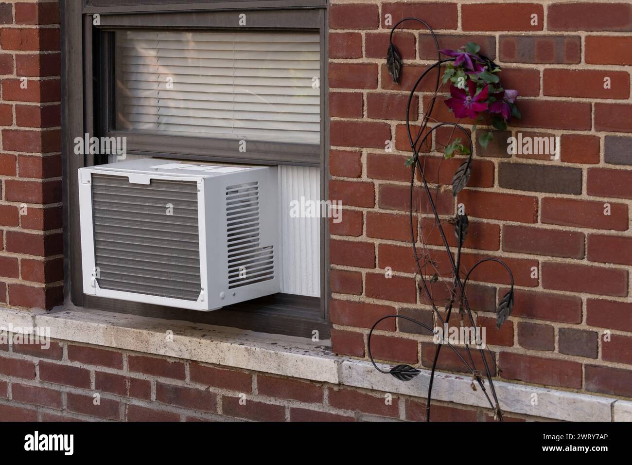 Außenansicht der Klimaanlage Fenstereinheit, die von der Fensterbank eines roten Backsteingebäudes mit einer deorativen Blume daneben ragt Stockfoto