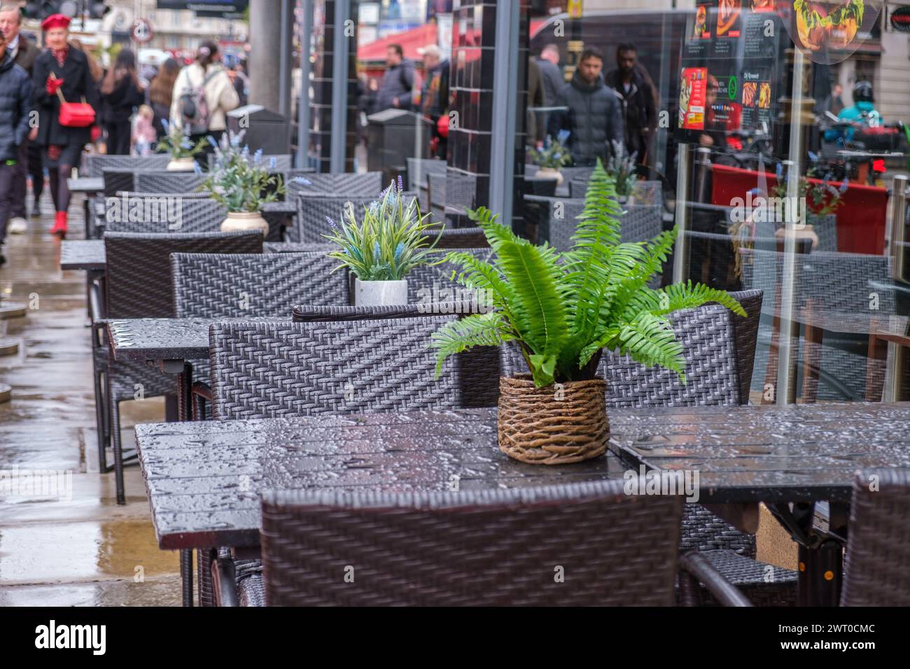 Regengetränktes Straßencafé mit nassen Tischen und Stühlen, Topfpflanzen und Fußgängern im unscharfen Hintergrund. Zentrum Von London, England. Stockfoto