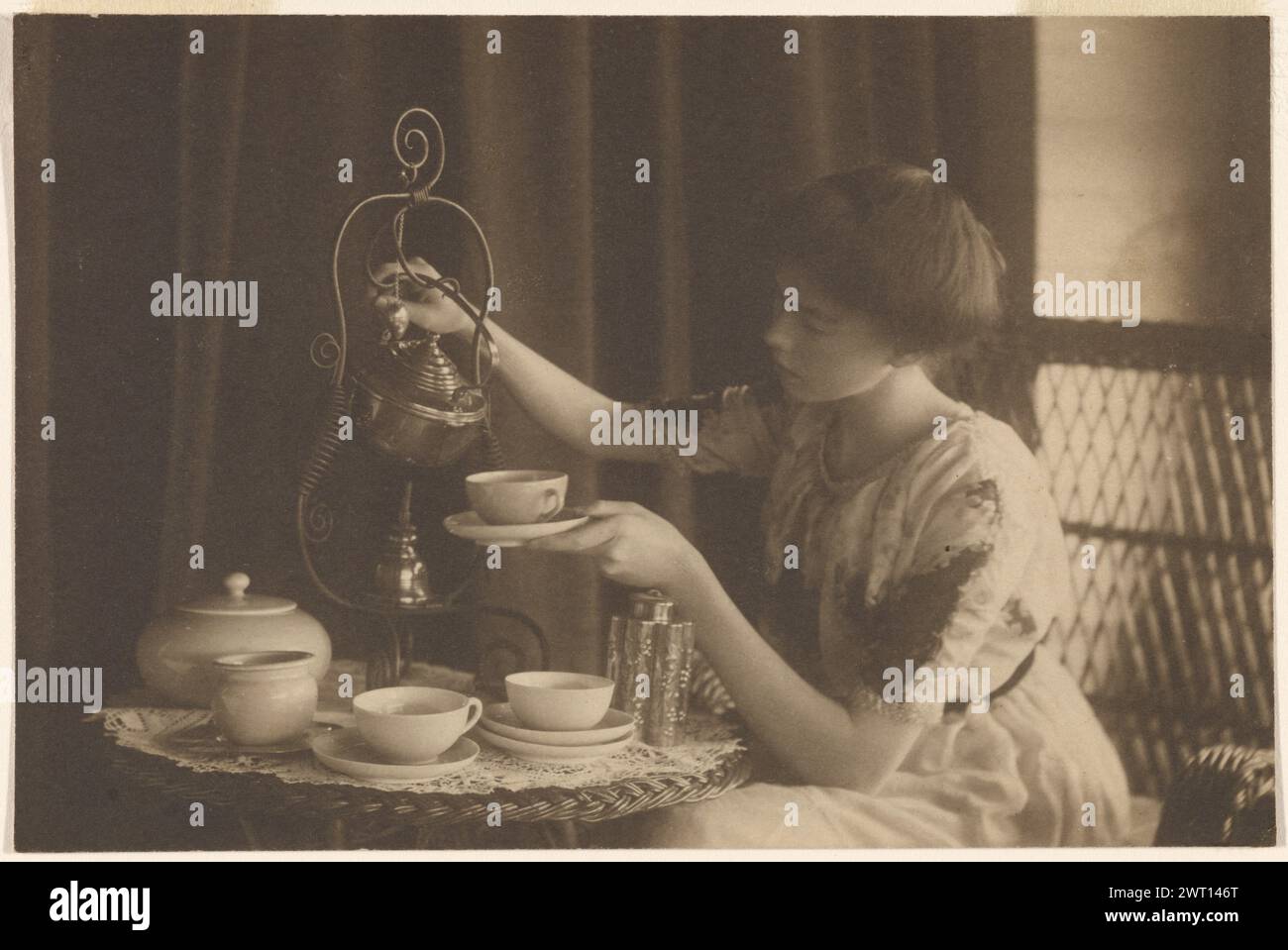 Frau, die Tee gießt. Alice Barber Stephens, Fotografin (Amerikanerin, 1858–1932) 1900–1920 Eine junge Frau, die an einem Tisch saß und eine Tasse Tee aus einer hängenden Teekanne goss. Auf dem Tisch stehen leere Tassen und Untertassen. (Rückseitig, bedruckt) obere Mitte, blaue Tinte: 'Jolen Walton'; (rückseitig bedruckt) Mitte rechts, schwarze Tinte: '[illeg] könnte'; (rückseitig bedruckt) unten links, Bleistift: '5/6'; Stockfoto