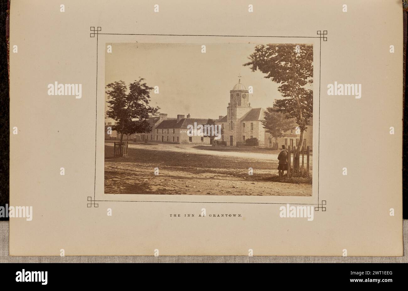 Das Inn in Grantown. George Washington Wilson, Fotograf (Schottisch, 1823 - 1893) 1868 Blick auf die Gebäude von Grantown-on-Spey entlang einer breiten Feldstraße, einschließlich des Grant Arms Hotel, dessen Name auf einer weißen Grenze in der Nähe der Dachlinie gemalt ist. Ein Mann steht neben einem Baum, umgeben von einem Zaun auf der rechten Seite des Bildes. (Recto, Mount) unten in der Mitte, Buchdruck in schwarzer Tinte: "DAS GASTHAUS IN GRANTOWN." Stockfoto