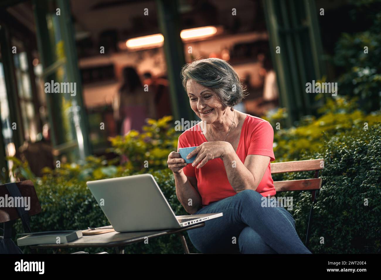 Ältere Frau, die im Café im Freien sitzt und einen Laptop benutzt und Kaffee trinkt. Stockfoto