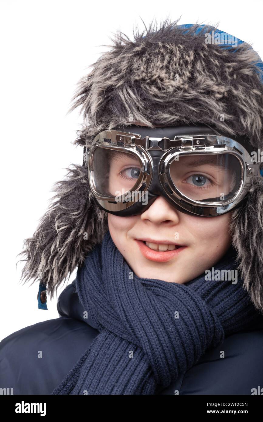 Lächelnder junger Junge, der für Winterspaß gekleidet ist, mit Pilotenbrille und Trapper-Hut aus Kunstfell vor weißem Hintergrund bereit Stockfoto