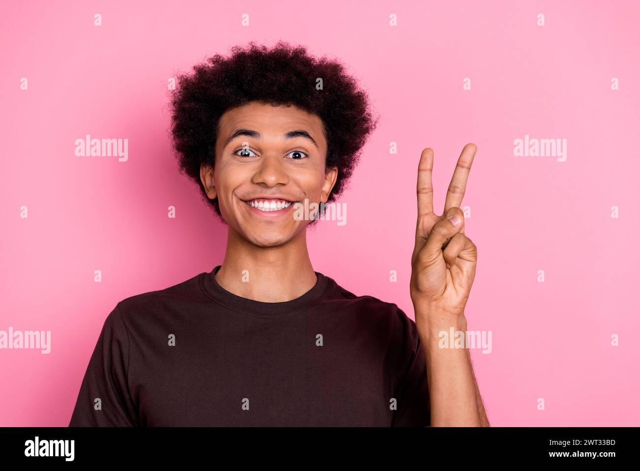 Porträt des jungen Lächelns fröhlicher Kerl in braunem T-Shirt zeigt V-Zeichen Hallo-Symbol Frieden gegen Krieg isoliert auf rosafarbenem Hintergrund Stockfoto