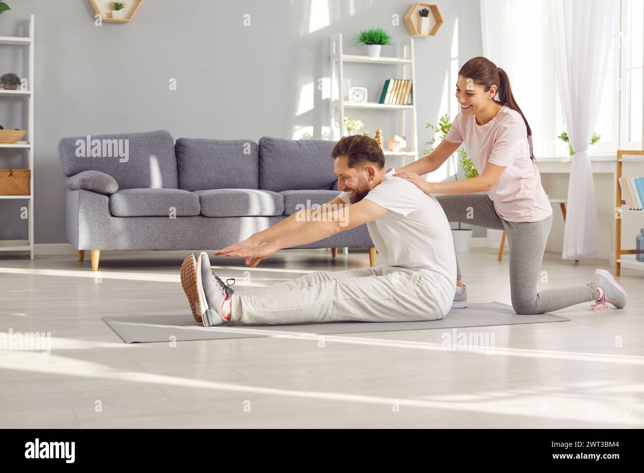 Glückliche, sportliche junge Frau, die ihrem Mann bei Sportübungen zu Hause hilft Stockfoto