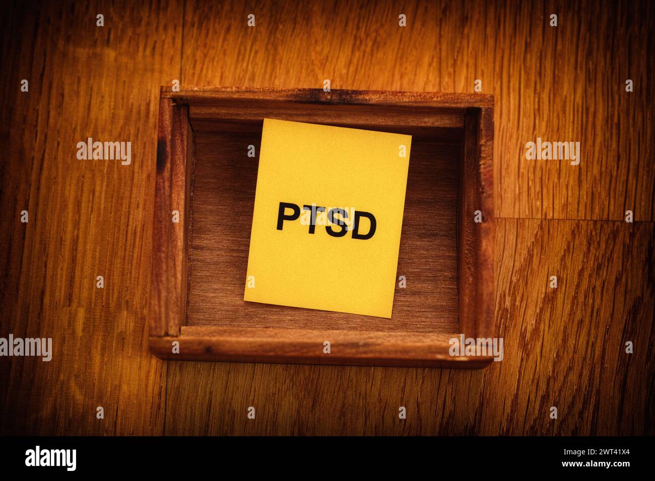 Ein gelbes Stück Papier mit der Abkürzung PTSD (posttraumatische Belastungsstörung), das in einer kleinen leeren Holzkiste liegt. Nahaufnahme. Stockfoto