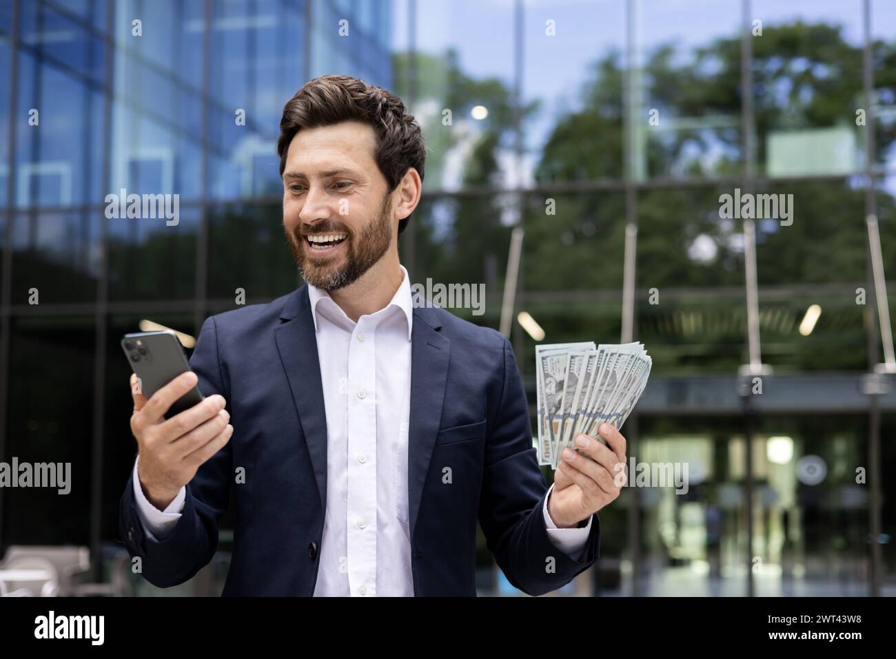 Glücklicher junger Mann, der einen Fan von Dollarscheine in der Hand hält und lächelnd auf den Bildschirm des Telefons blickt, in einem Idol-Anzug in der Nähe des Bürozentrums. Stockfoto