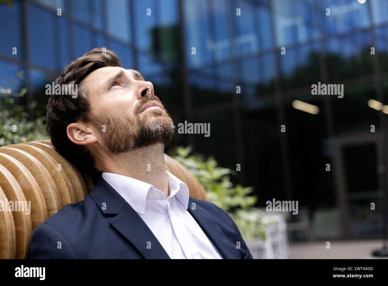 Nahaufnahme eines jungen nachdenklichen und müden Geschäftsmannes, der vor einem Bürogebäude sitzt, sich auf eine Bank lehnt, aufblickt, aufgeregt und enttäuscht. Stockfoto