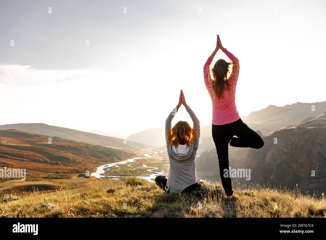 Zwei junge Frauen entspannen sich in Yoga-Posen in den Bergen und am Fluss bei Sonnenuntergang Stockfoto
