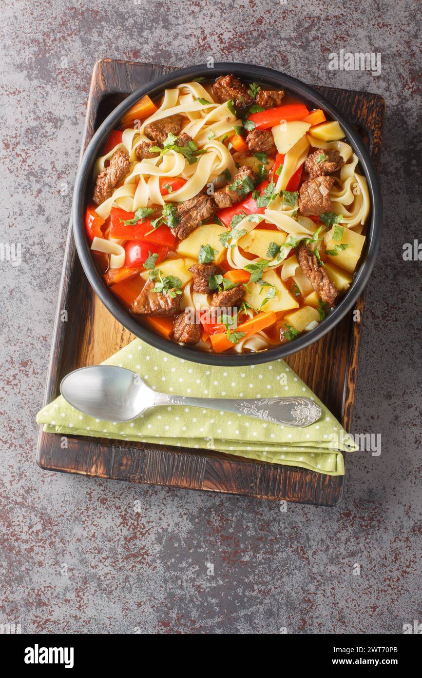 Lagman-Suppe mit Rindfleisch, Nudeln, Paprika, Kartoffeln, Karotten, Zwiebeln und Kräuter Nahaufnahme auf dem Teller auf dem Holzbrett. Vertikale Draufsicht von oben Stockfoto