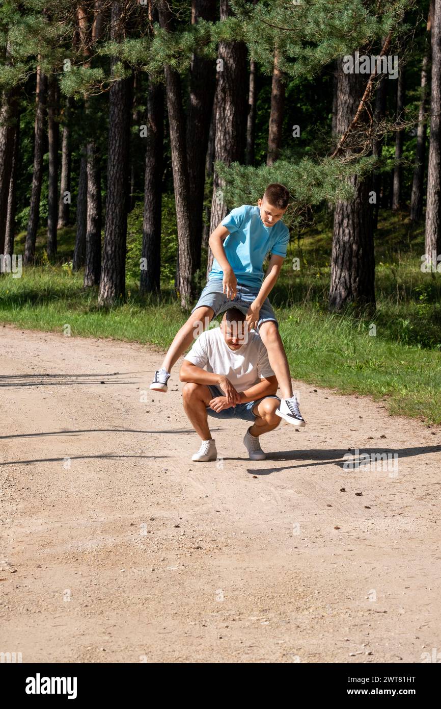 Ein Vater hockt sich, als sein Sohn über den Rücken springt, beide genießen einen sonnigen Tag in einem Kiefernwald, der für Familienverbundenzeit in der Natur genutzt wird. Hochwertige Pho Stockfoto