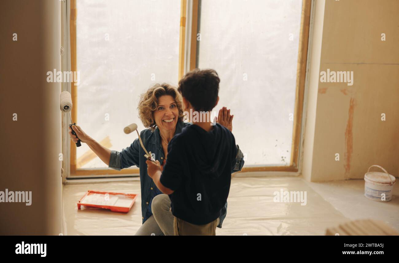 Mom und ihr Sohn feiern mit High Five, während sie während einer Hausrenovierung eine Wand zusammen malen. Ihr Lächeln spiegelt ihr Glück wider und zeigt das Stockfoto