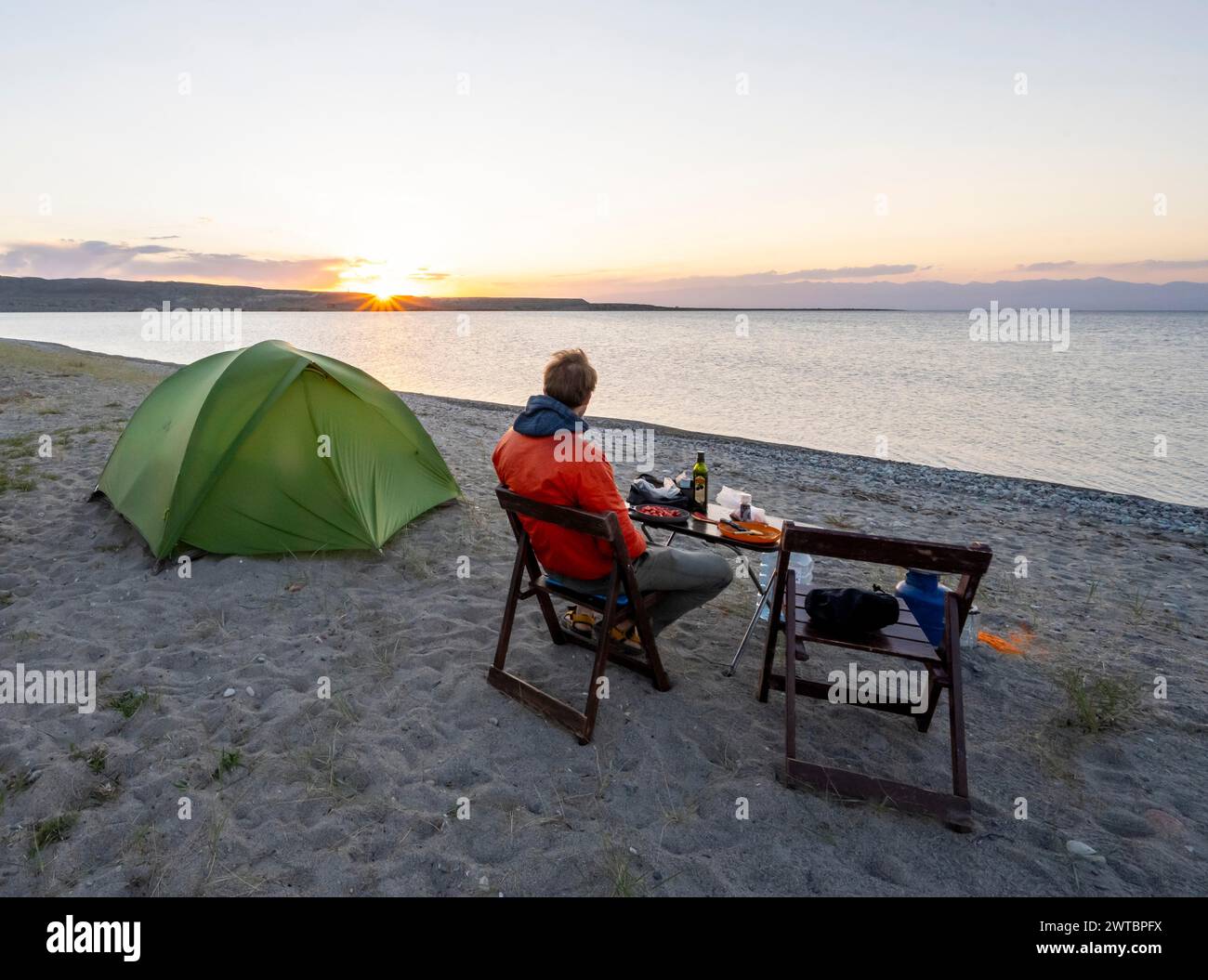 Camping am Seeufer, junger Mann sitzt beim Abendessen auf einem Campingstuhl neben dem Zelt, Sonnenuntergang am Issyk Kul See, Issyk Kul, Kirgisistan Stockfoto