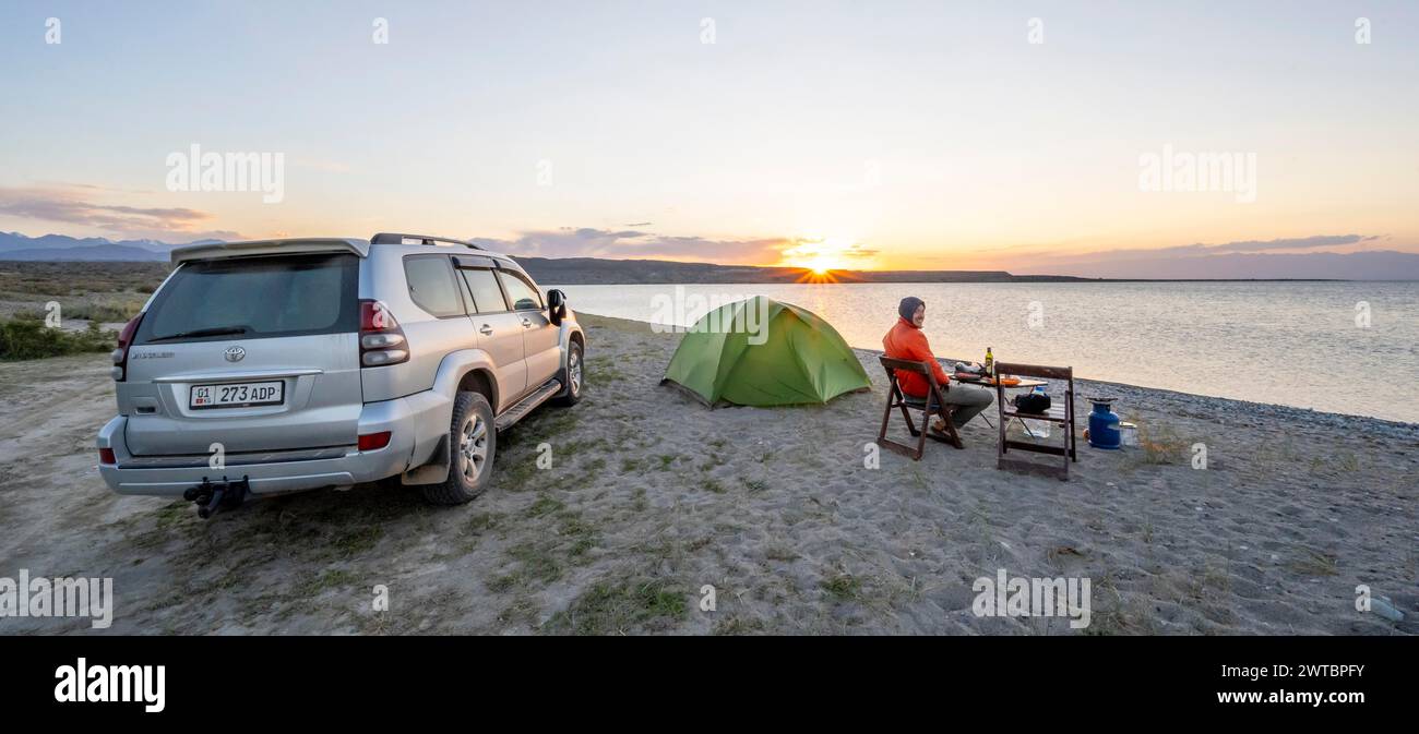 Camping am Seeufer, junger Mann sitzt neben dem Zelt auf einem Campingstuhl, Abendessen, Geländewagen, Sonnenuntergang am Issyk Kul See, Issyk Kul Stockfoto