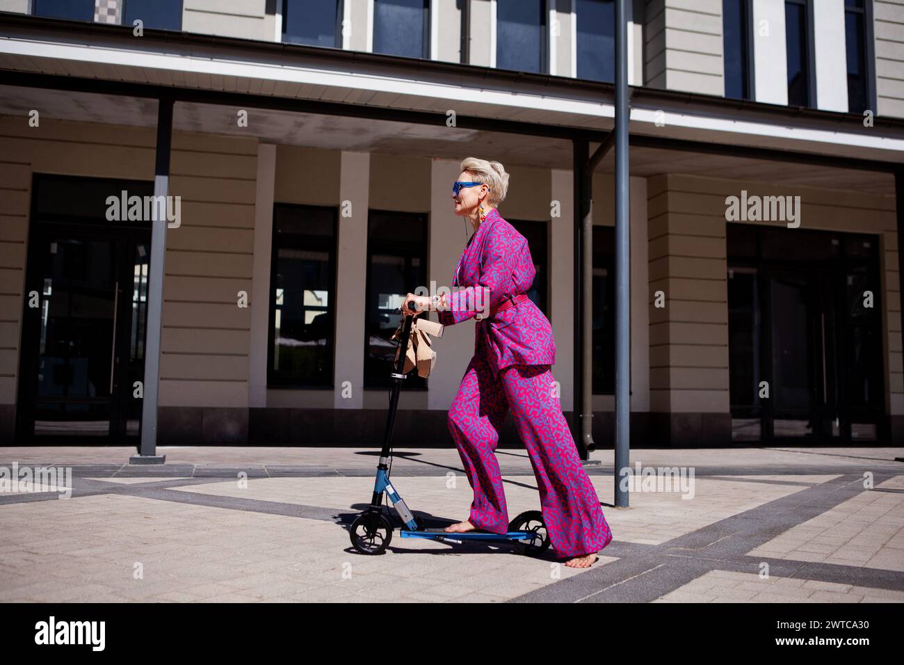 Stilvoll gekleidete reife Frau in einem knalligen pinken Hosenanzug und einer Jacke reitet mit einem Roller durch die Stadt, einem aktiven, gesunden und umweltfreundlichen Frien Stockfoto