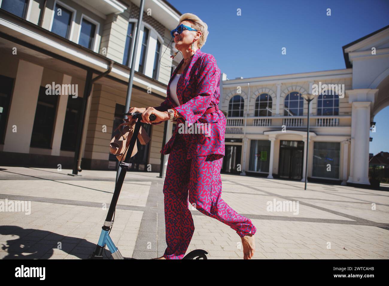 Stilvoll gekleidete reife Frau in einem knalligen pinken Hosenanzug und einer Jacke reitet mit einem Roller durch die Stadt, einem aktiven, gesunden und umweltfreundlichen Frien Stockfoto