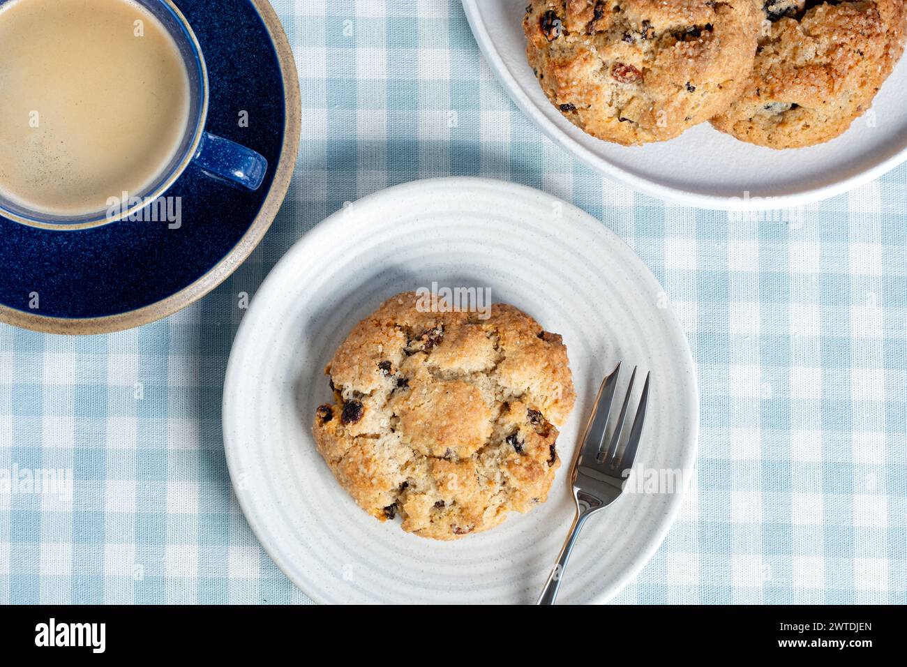 Ein frischer traditioneller, großer englischer Rock Cake serviert mit einer Tasse schwarzen Kaffee. Die Kuchen werden mit einer Gebäckgabel auf einem karierten Tischtuch überzogen Stockfoto