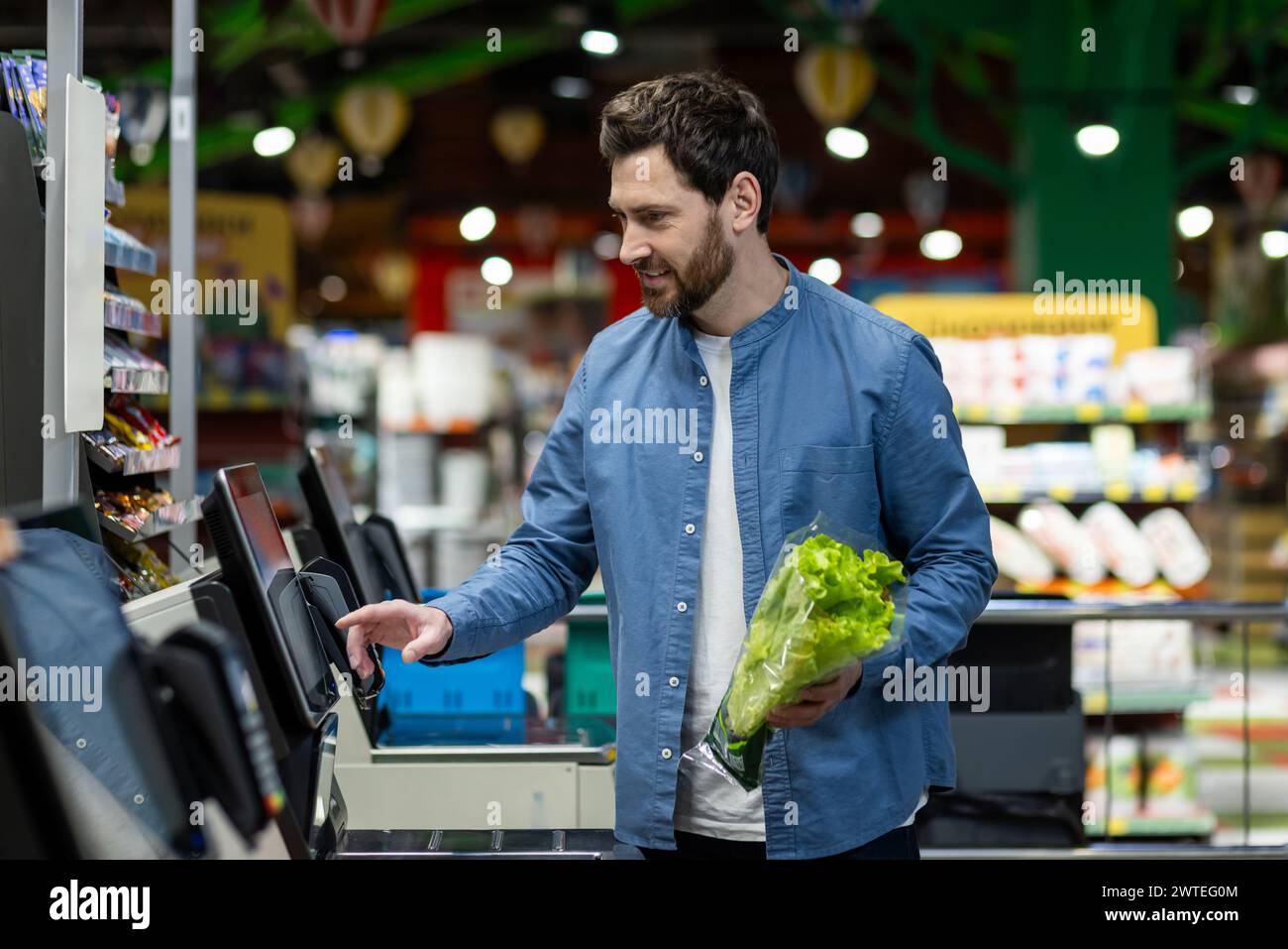 Erwachsener Mann in einem blauen Hemd mit einem Selbstkassenautomaten in einem Supermarkt, der frischen Salat hält. Stockfoto